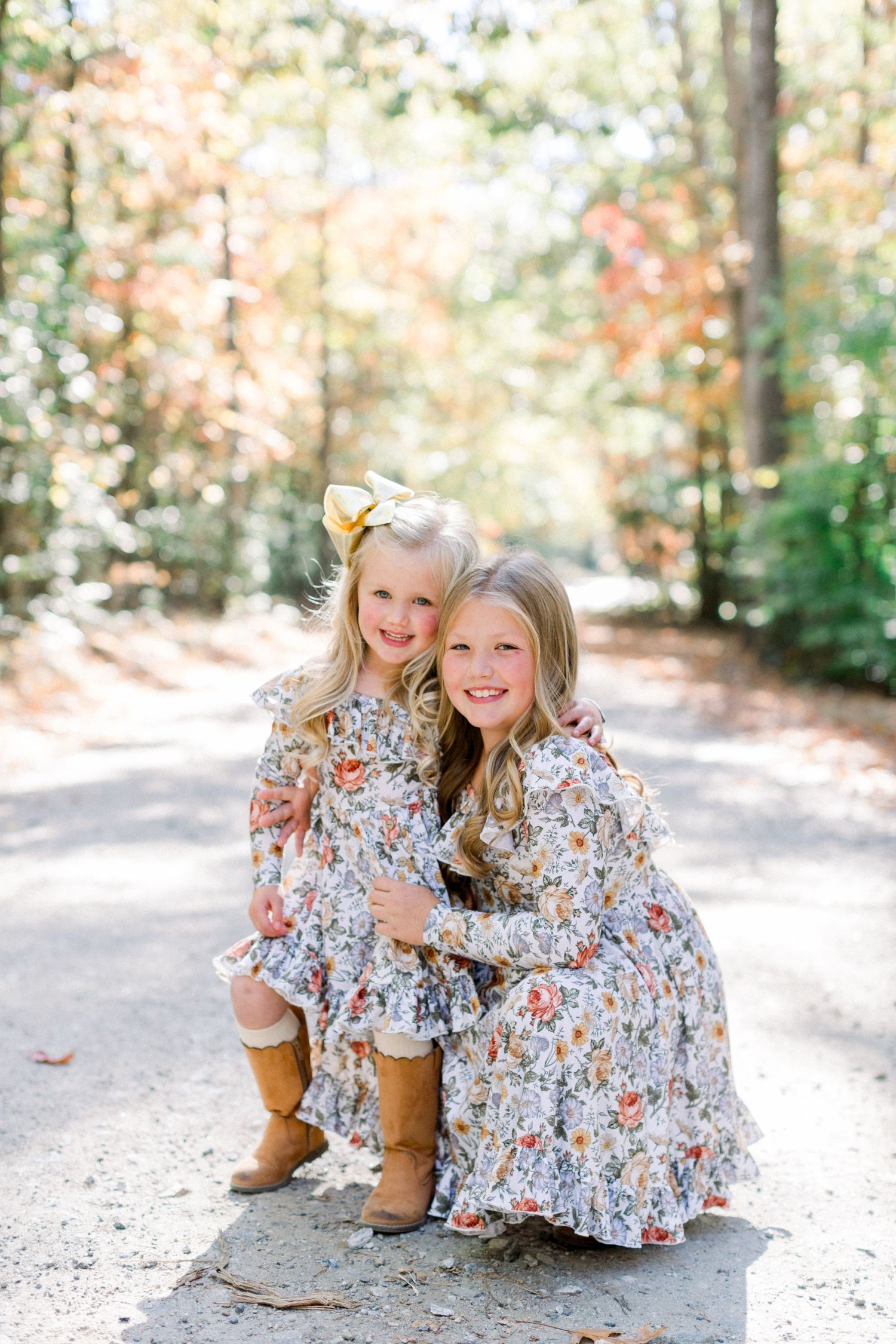 Two young sisters in matching floral dresses hugging on a sunlit path surrounded by fall foliage.