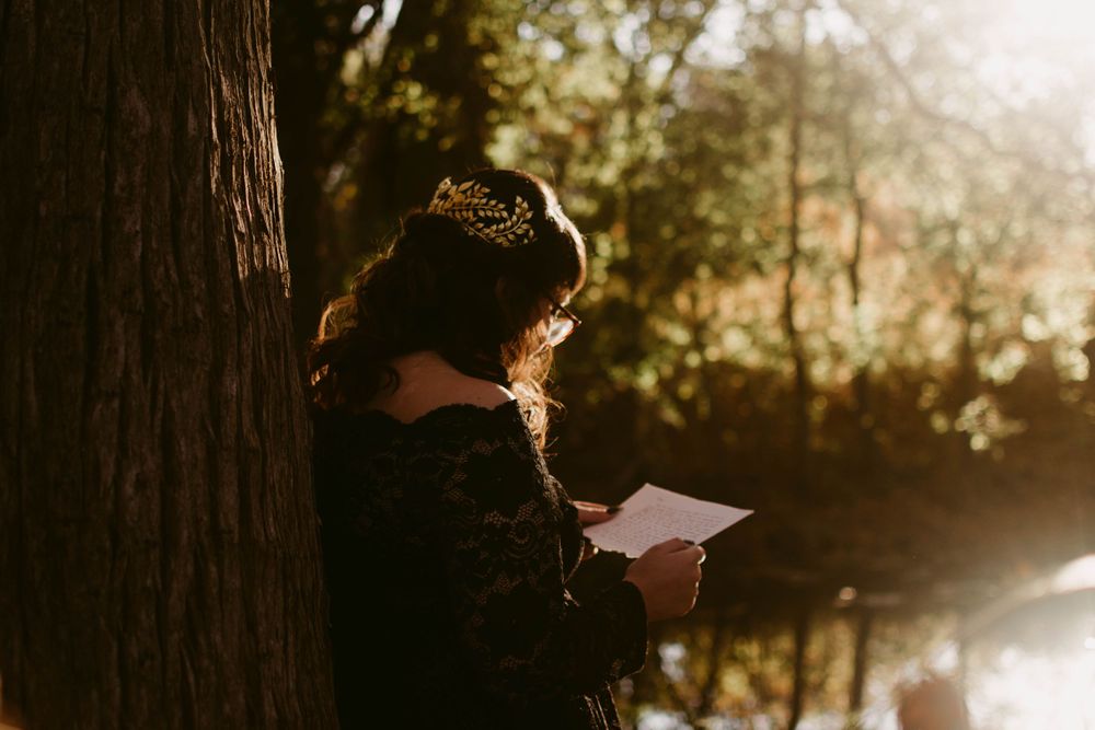Figure in dark clothing reads papers while resting against tree trunk in glowing forest light.