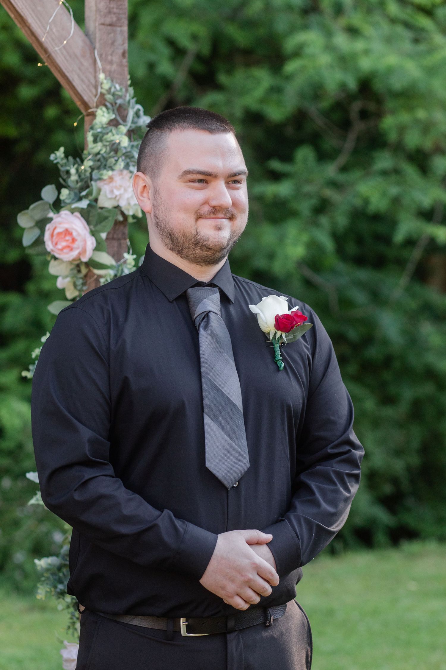 Groomsman in black suit and gray tie smiling during outdoor wedding ceremony.