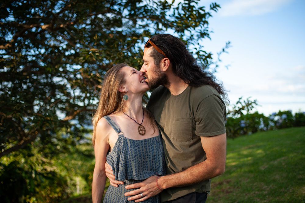 Amy biting Carey Primeau's lip on a green hill top in Costa Rica with trees and blue sky in the background.