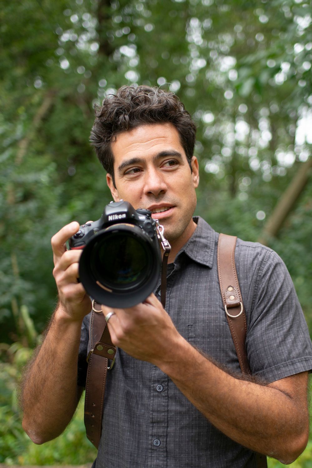 A photographer in a grey shirt holds a professional camera with a large lens in a forest setting.