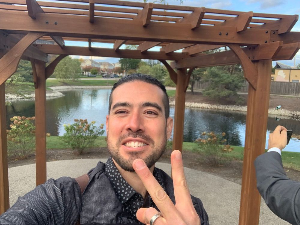 A selfie taken under a wooden pergola with a peaceful pond view in the background.