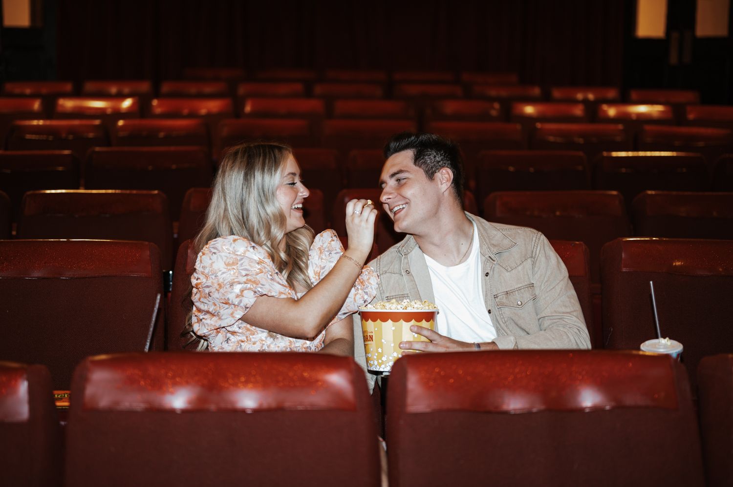 A couple sharing popcorn and laughing together in empty theater seats.
