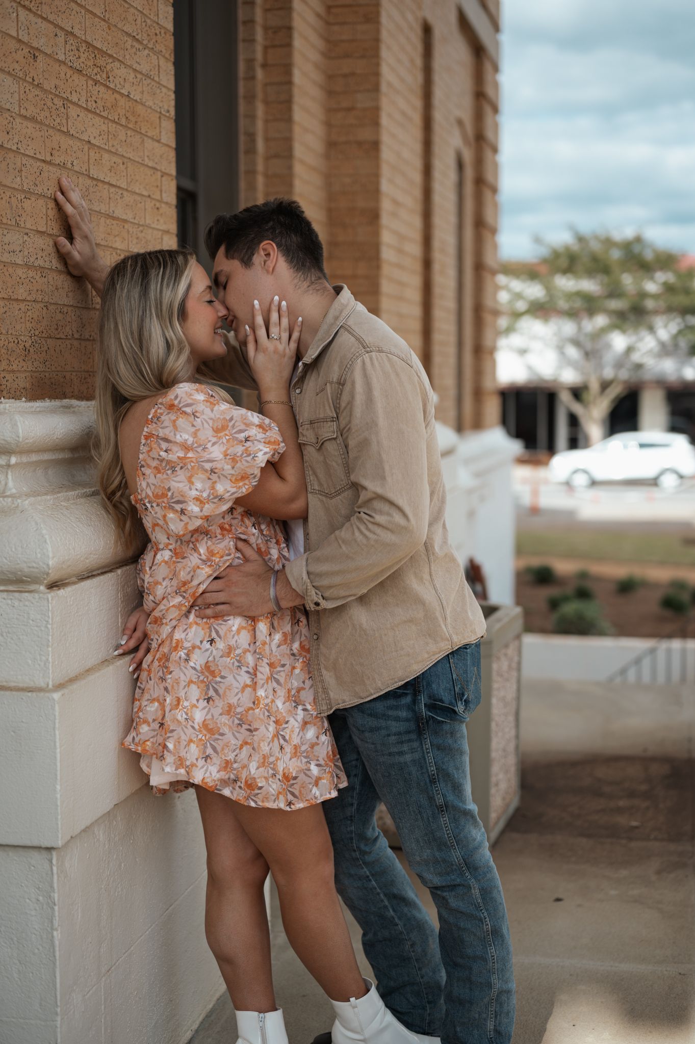 A couple in casual attire sharing an intimate moment against a brick wall exterior.
