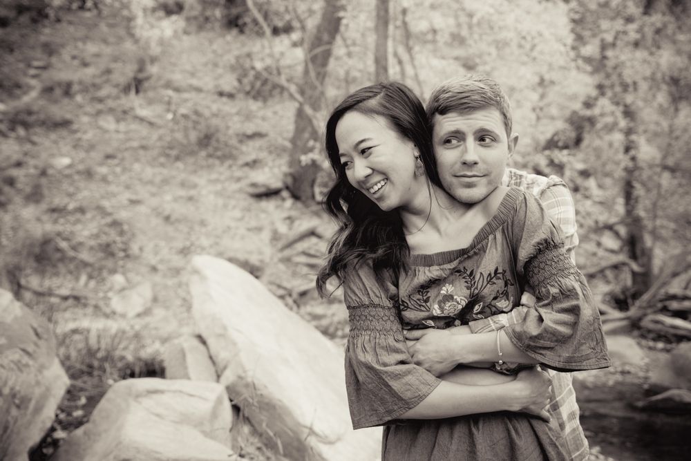 Black and white photo of a smiling couple embracing outdoors among rocks and trees.