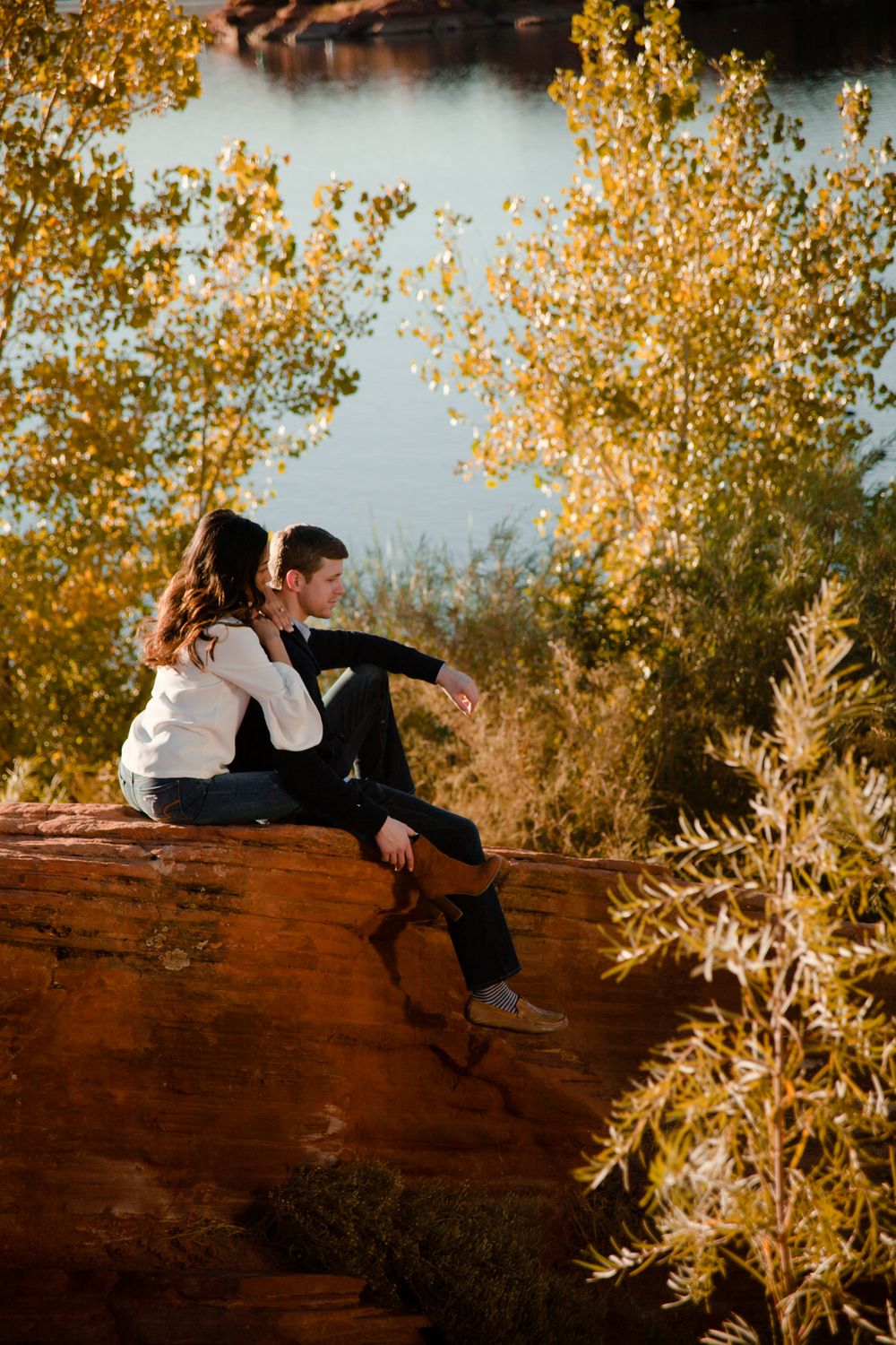Couple sitting on red rocks surrounded by golden autumn trees beside a tranquil lake.