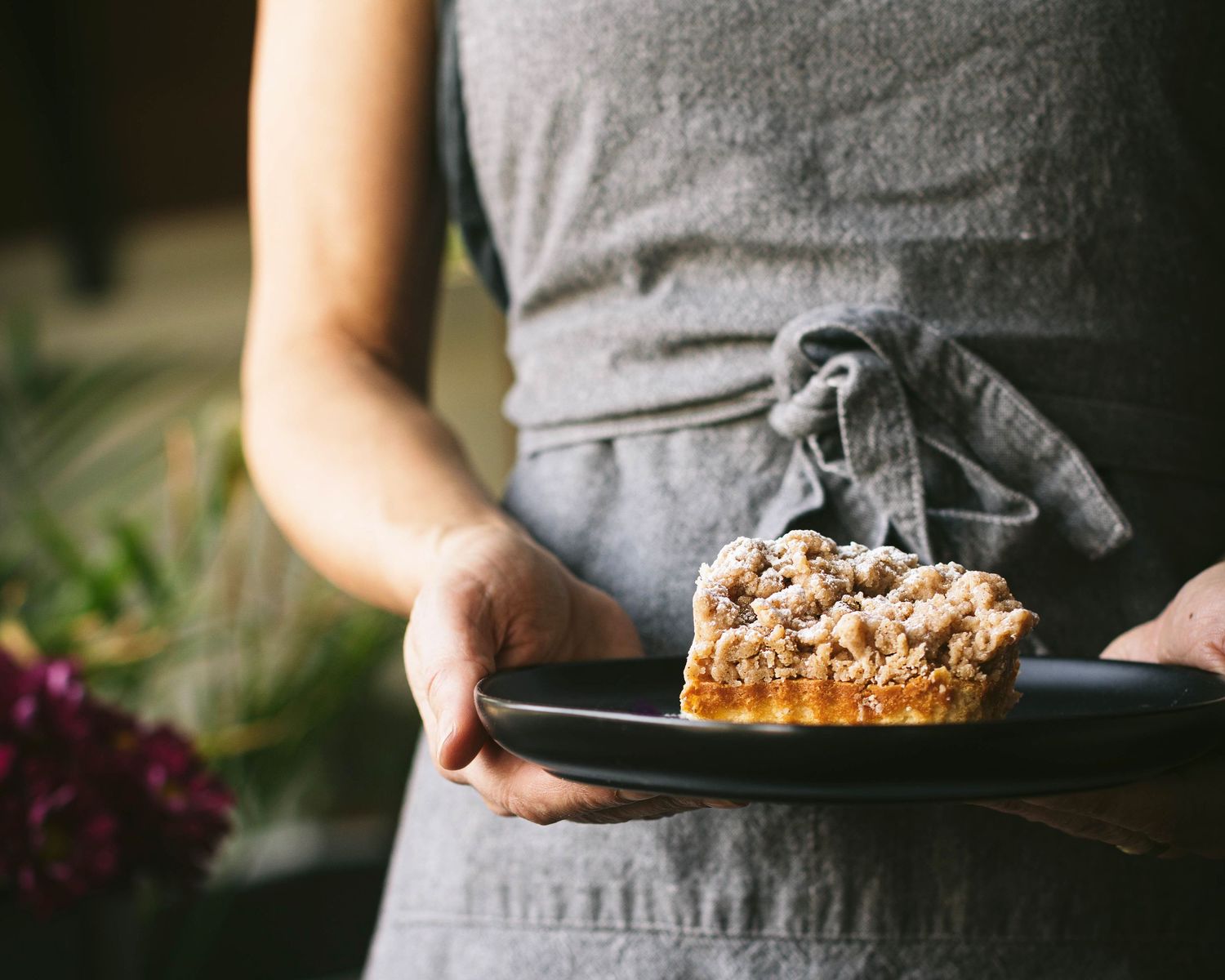 Person in gray apron holding a black plate with a rustic crumble-topped pastry slice.