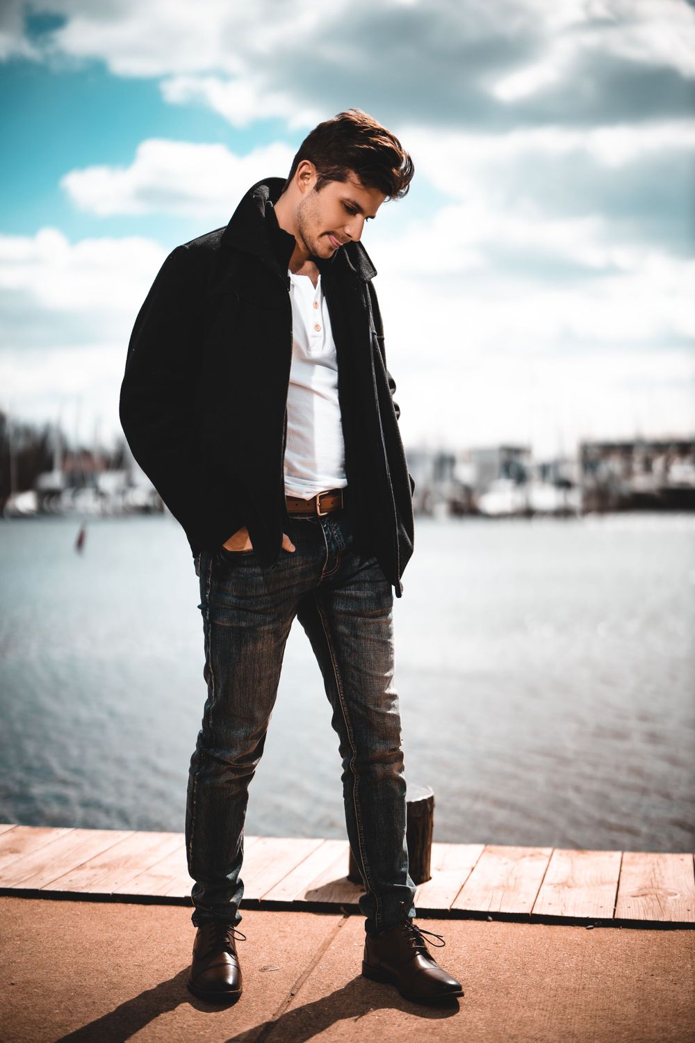 Fashion photo of a person in black jacket by a waterfront dock with moody sky.