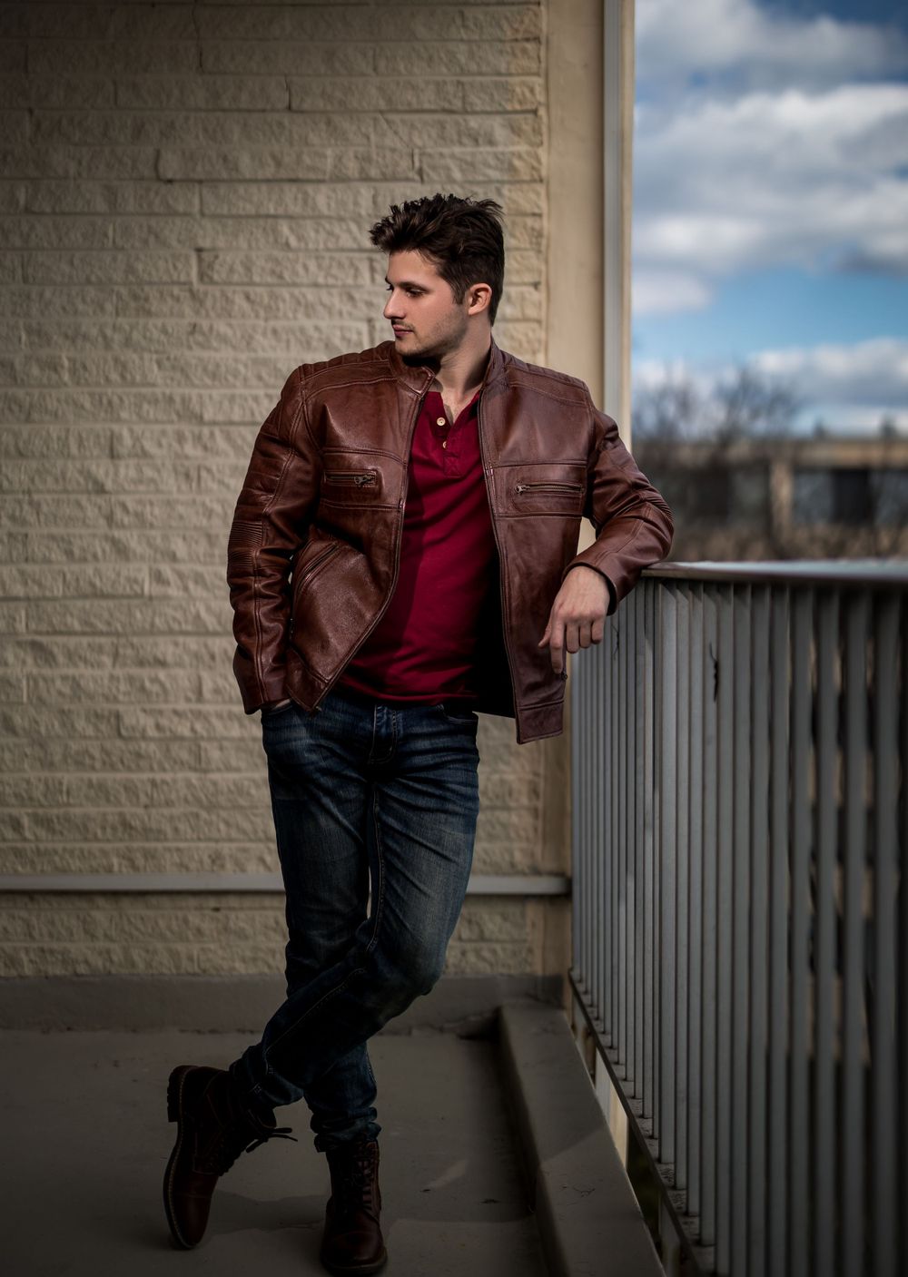 Fashion model in brown leather jacket and red henley poses against white brick wall.