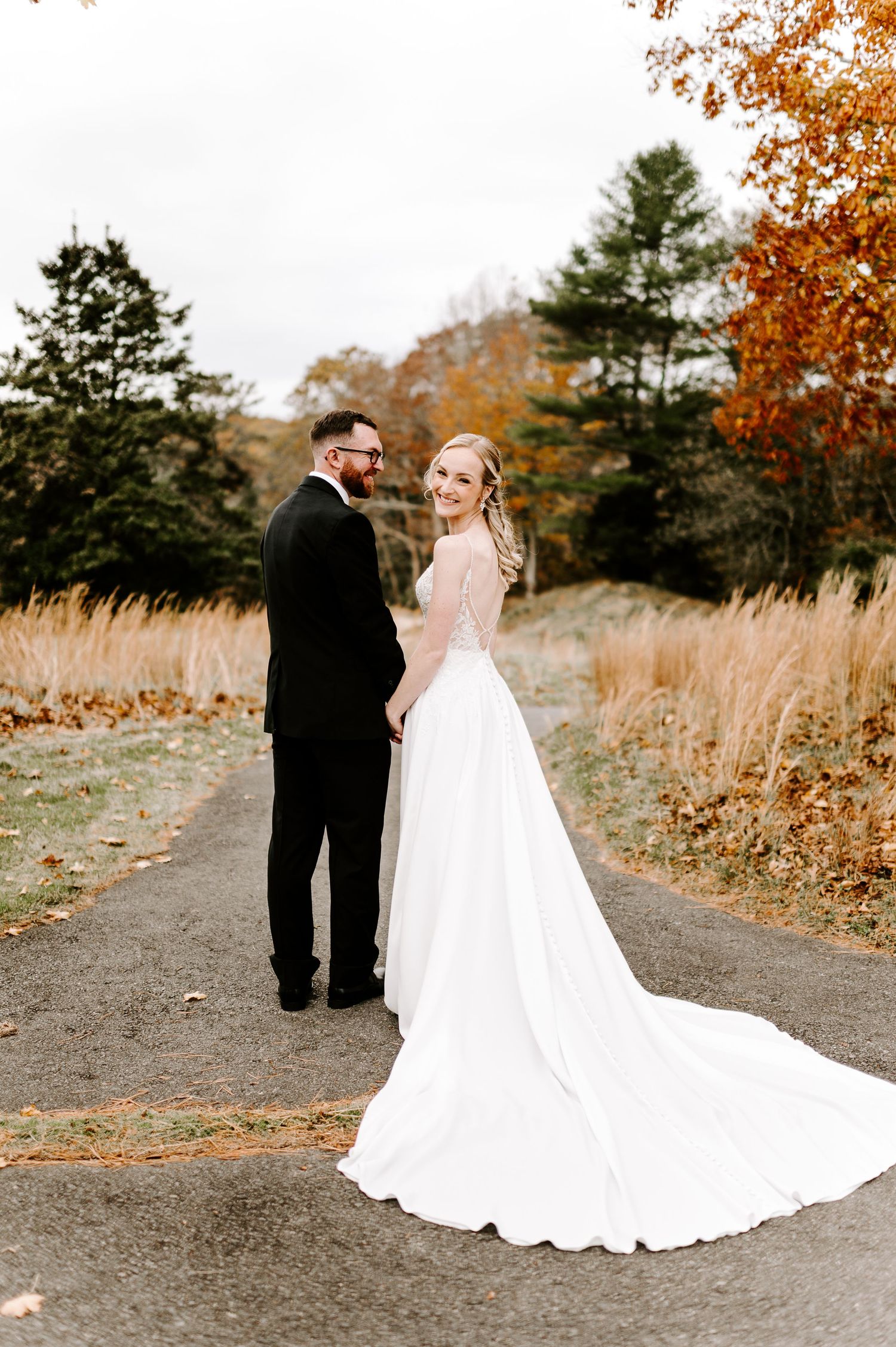 Wedding couple walking along gravel path surrounded by fall colors and tall grass.