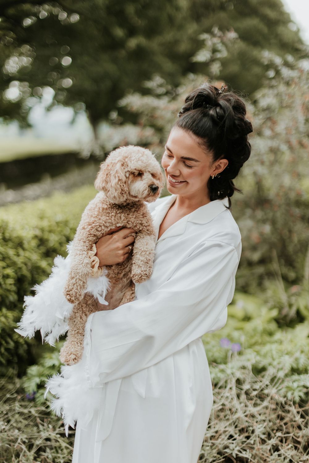 A person in white robe holds a fluffy cream-colored poodle mix dog in a garden setting.