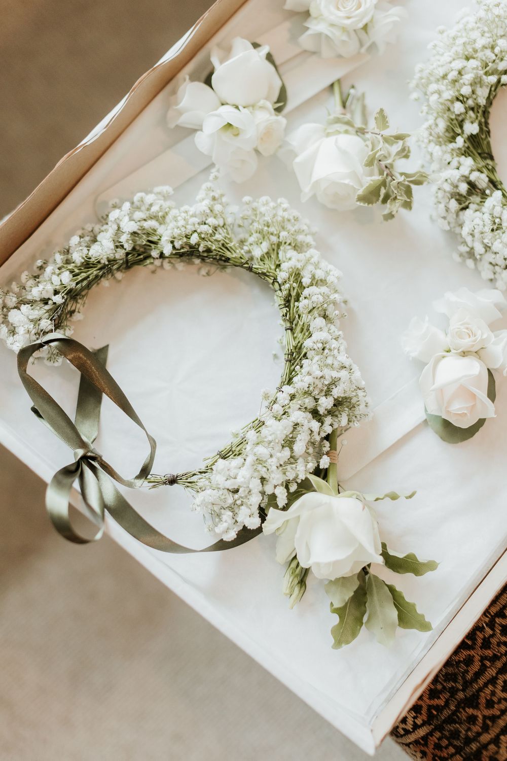 A delicate flower crown made of white baby's breath and roses with a gray ribbon lying on a white surface.
