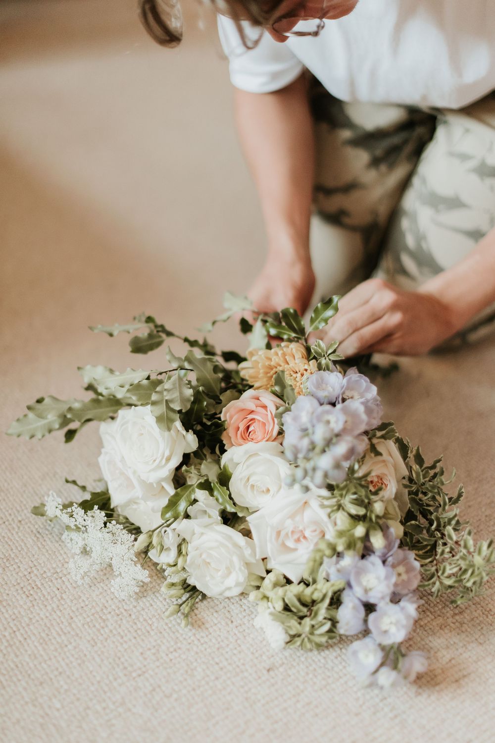 Hands arranging a romantic bouquet with white roses, peach flowers and purple accents.