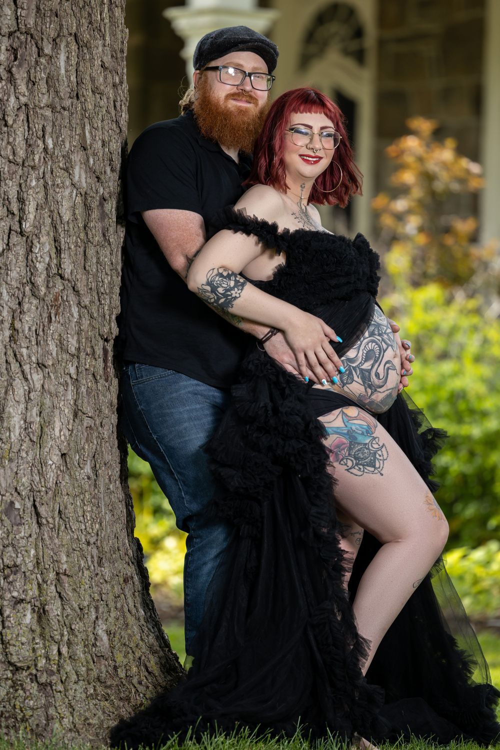 A couple in gothic attire posing intimately against a tree trunk in a garden setting.