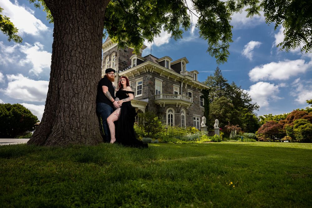 Couple poses beneath large tree with historic mansion in background.