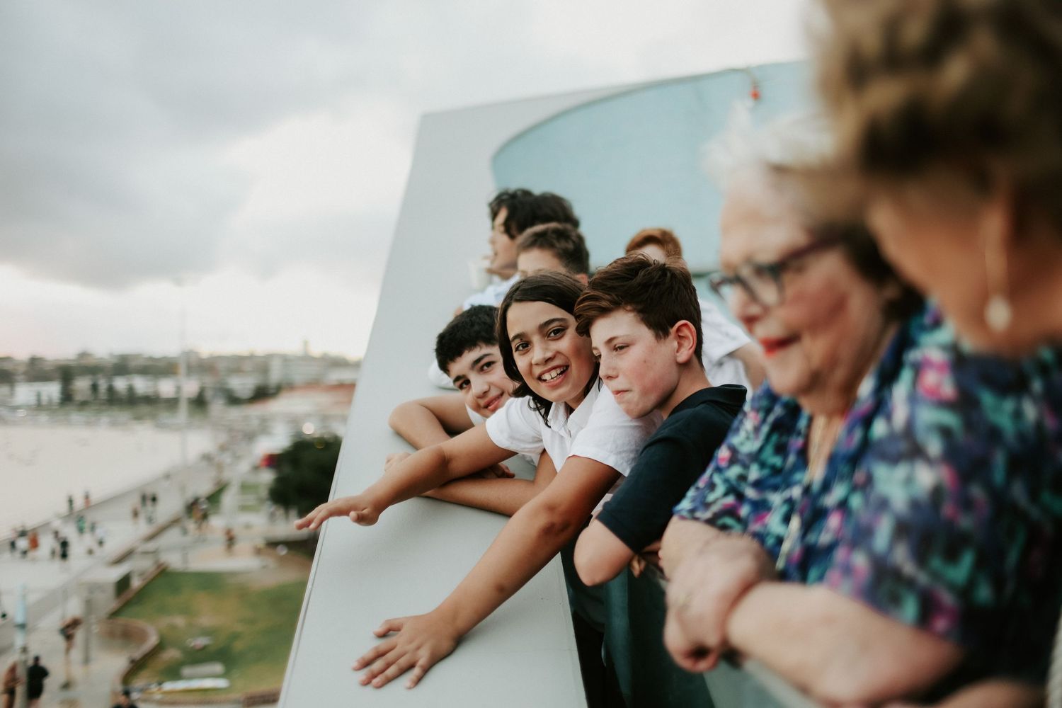 Group of smiling children and elderly woman leaning over railing at waterfront.