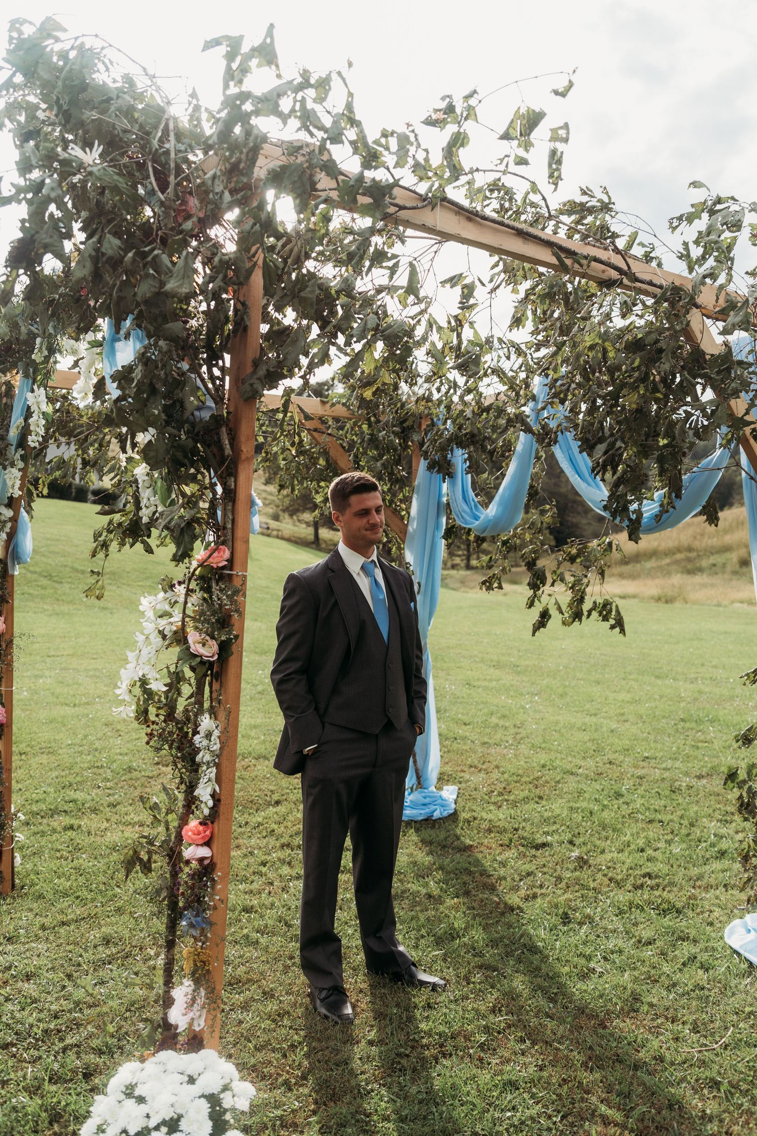A wedding ceremony setup outdoors with wooden posts draped in light blue fabric and greenery in a grassy field.