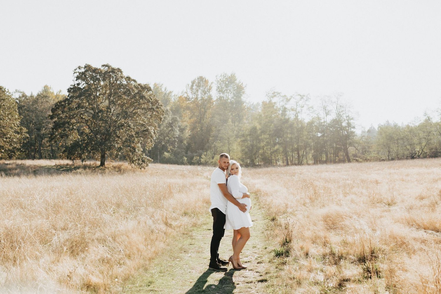 A couple embraces in a golden sunlit field with tall grass and trees in the background.