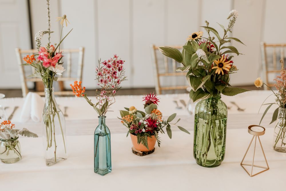 Mixed vintage bottles and geometric vases filled with colorful wildflowers on white tablecloth.