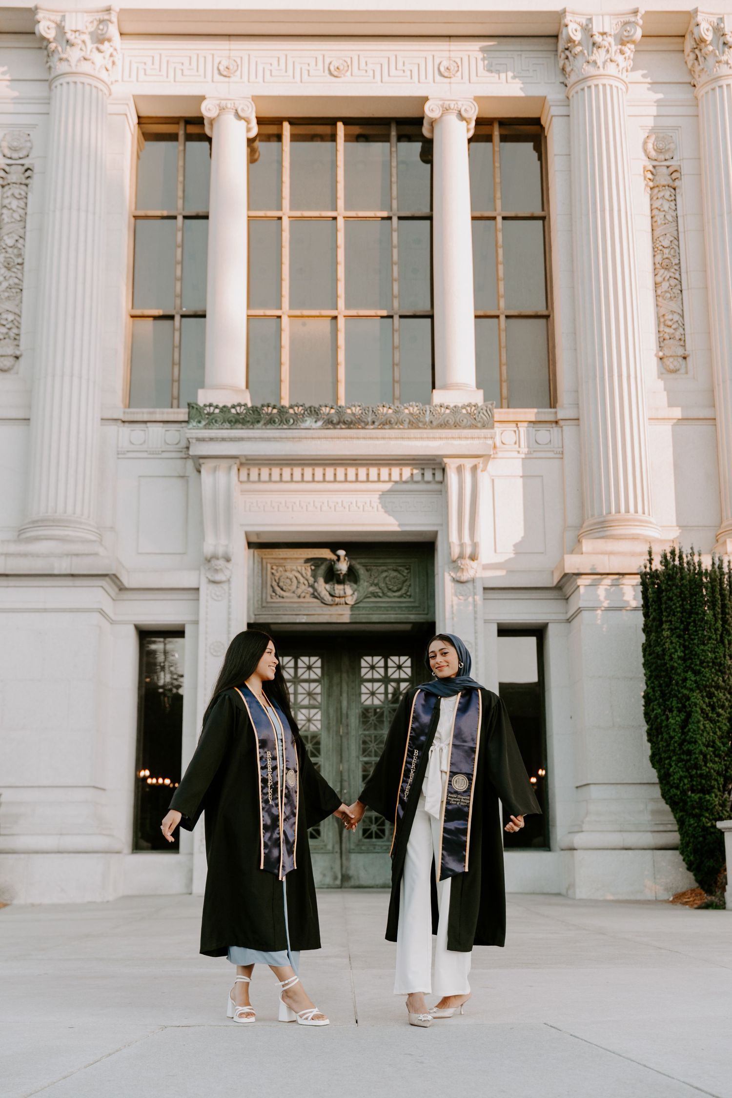 Two graduates in black robes hold hands in front of ornate university building entrance.