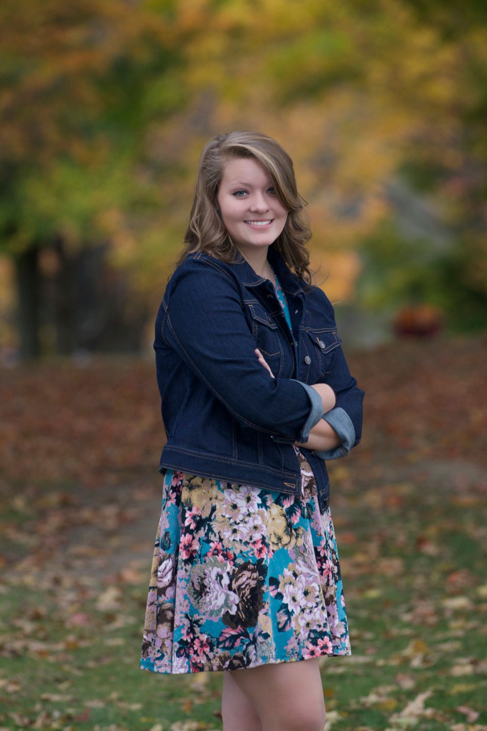 Person in denim jacket and floral dress poses against fall foliage background.