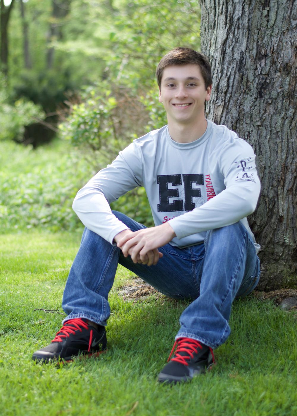 Student in light gray sweatshirt and jeans sits casually against large tree trunk.