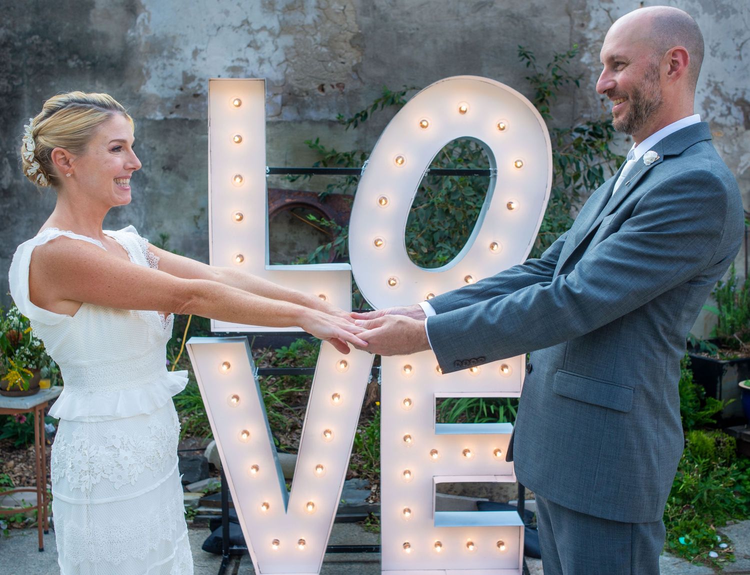 Newlyweds pose with a large illuminated LOVE sign at their outdoor wedding reception.