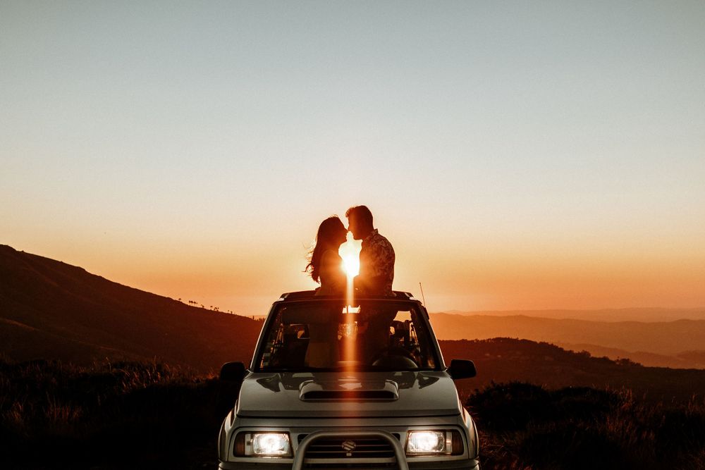 Couple silhouetted kissing on top of SUV at breathtaking mountain sunset with golden sky backdrop.