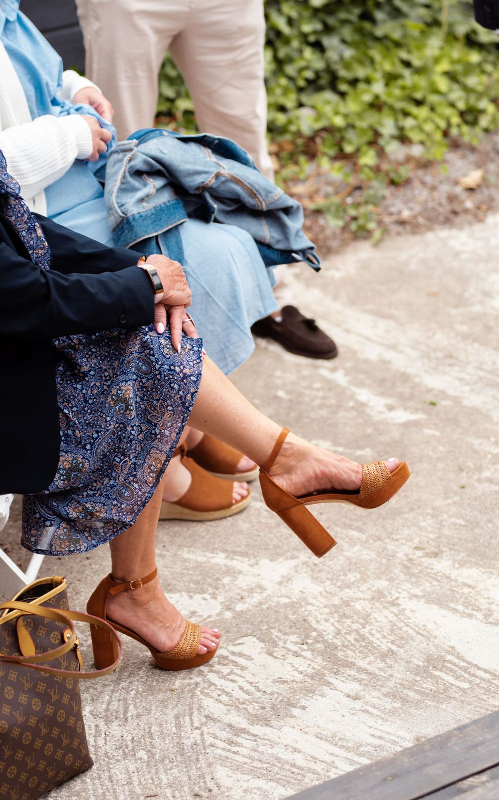 Women's feet in brown heels sitting on concrete steps with bags and clothing visible.