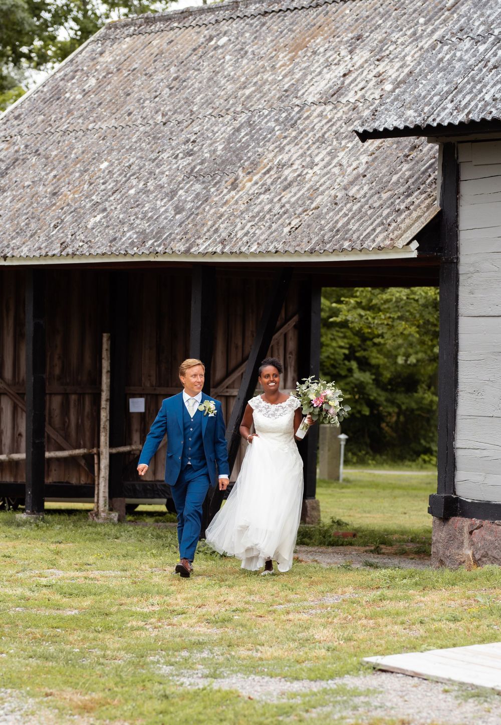 Two wedding guests walk together while exiting a rustic barn with weathered wood and shingle roof.
