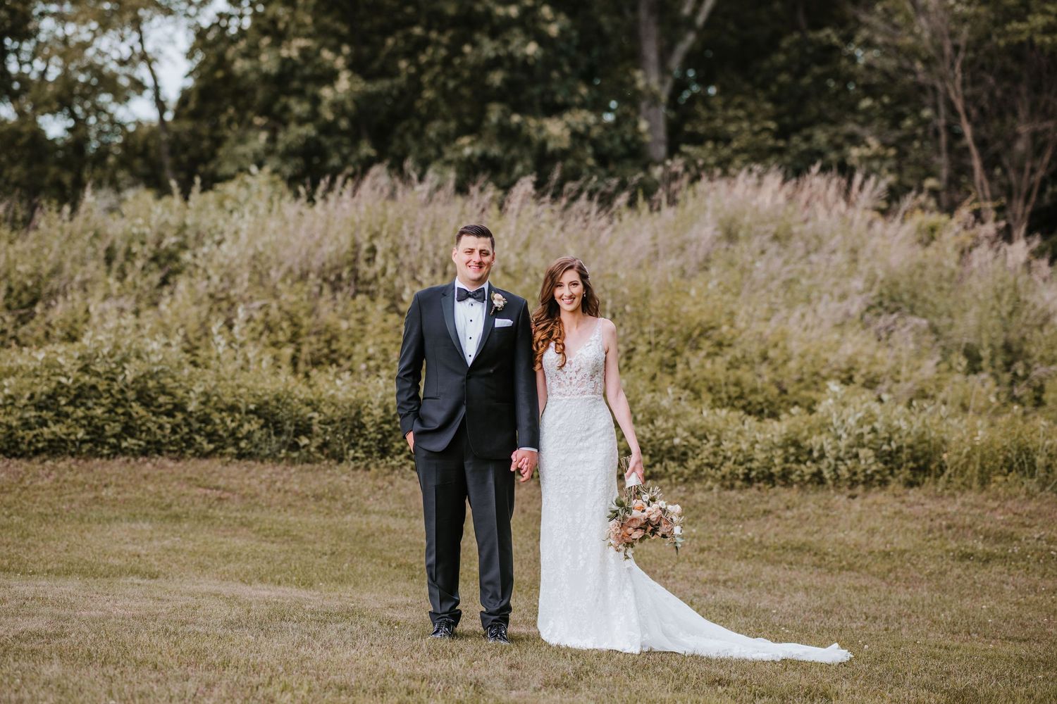 A newlywed couple stands together in a meadow with tall grass and wildflowers in the background.