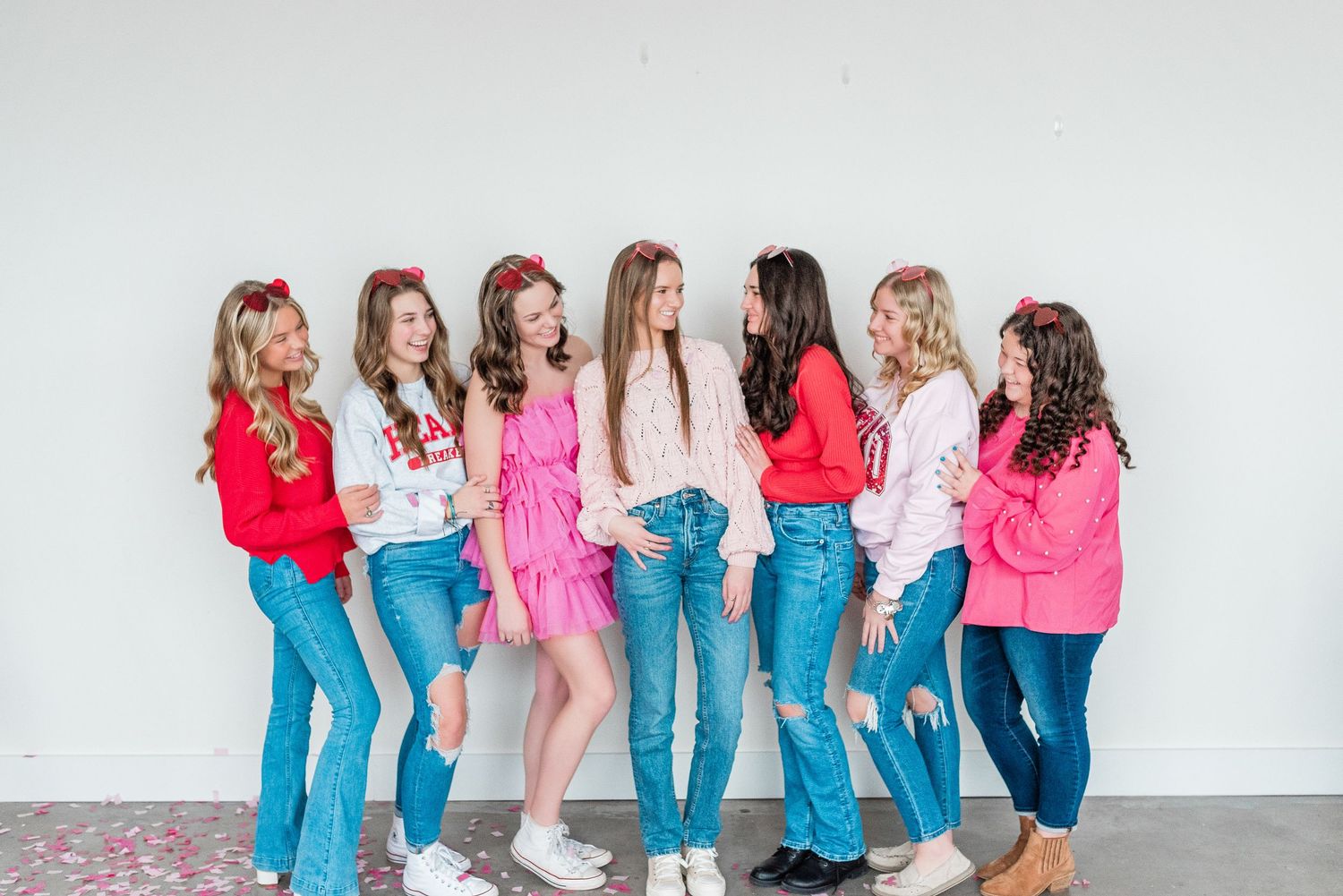 Group of friends in coordinating pink tops and blue jeans lean against white wall in casual poses.