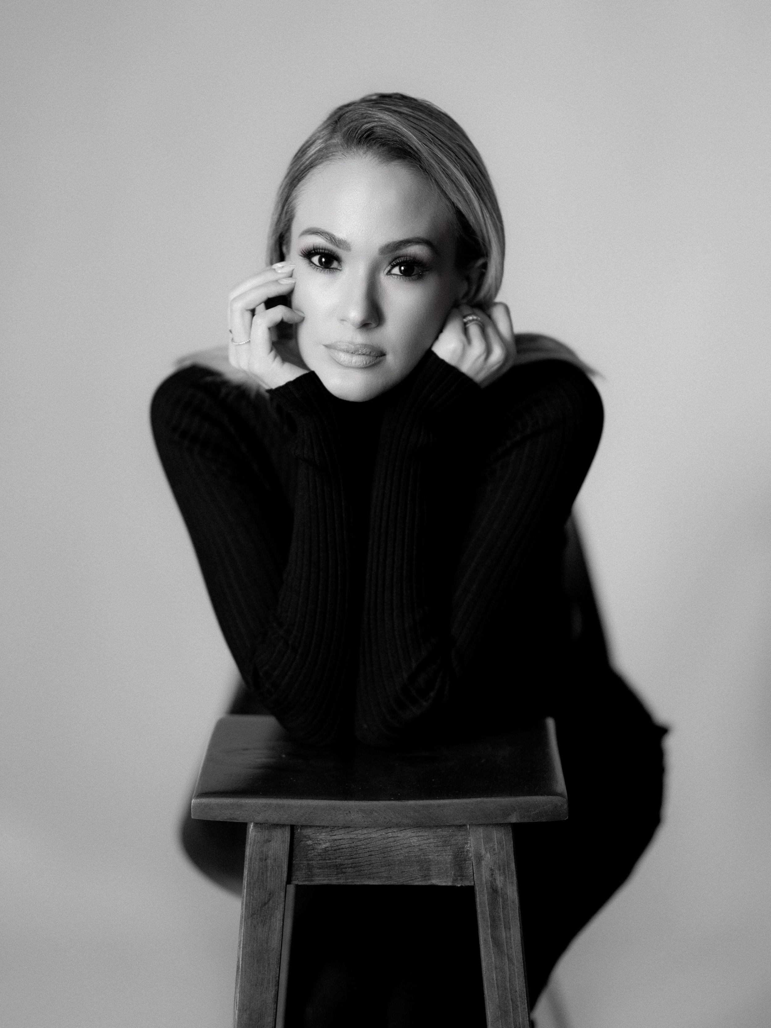 Professional studio portrait showing elegant pose on wooden stool against light background wearing black sweater.