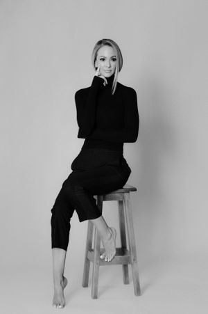 Artistic monochrome portrait showing relaxed pose on wooden stool in all-black ensemble.