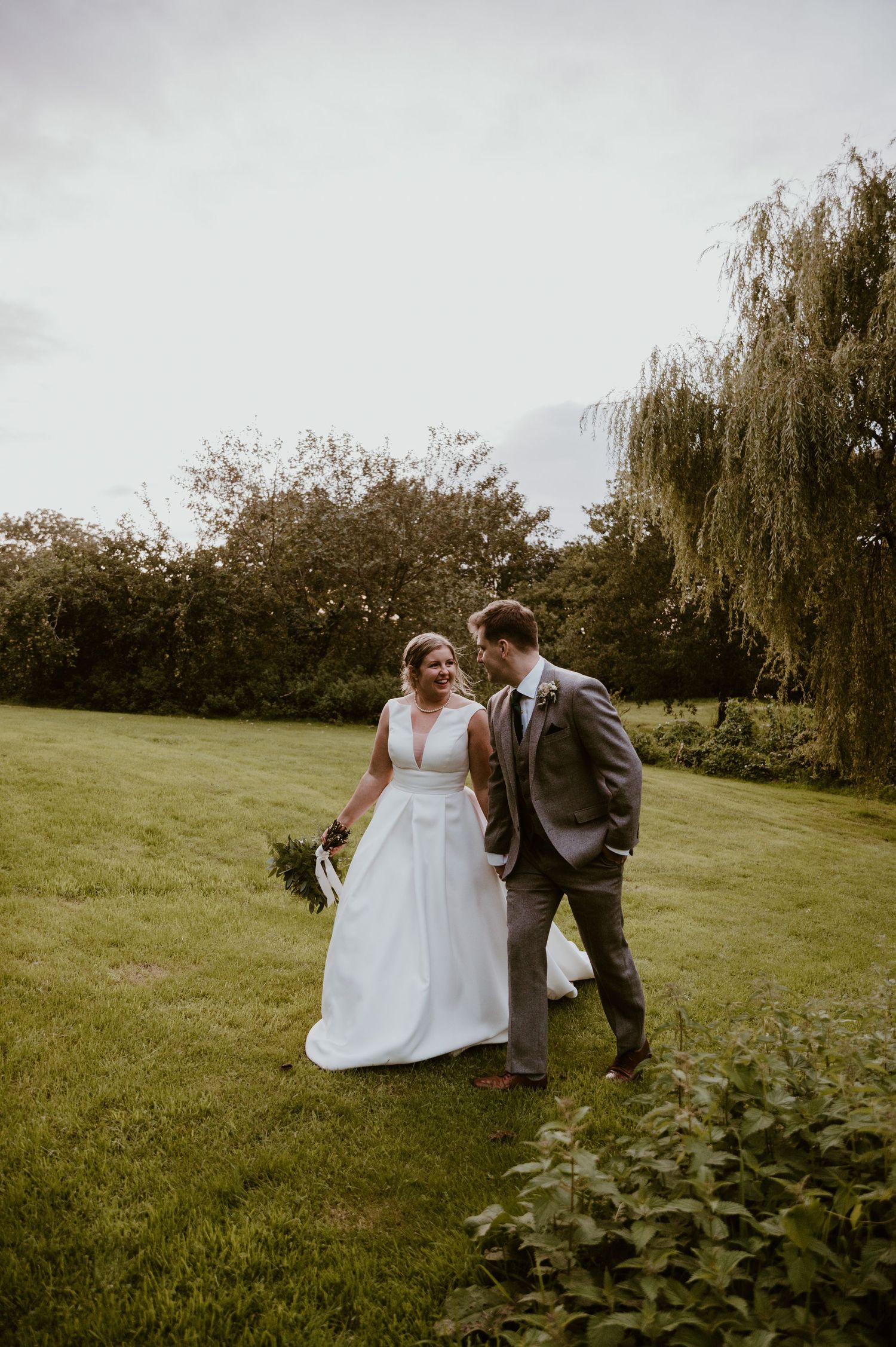 A romantic wedding moment captured on a lush green lawn with a willow tree and overcast sky in the background at Hareston Manor