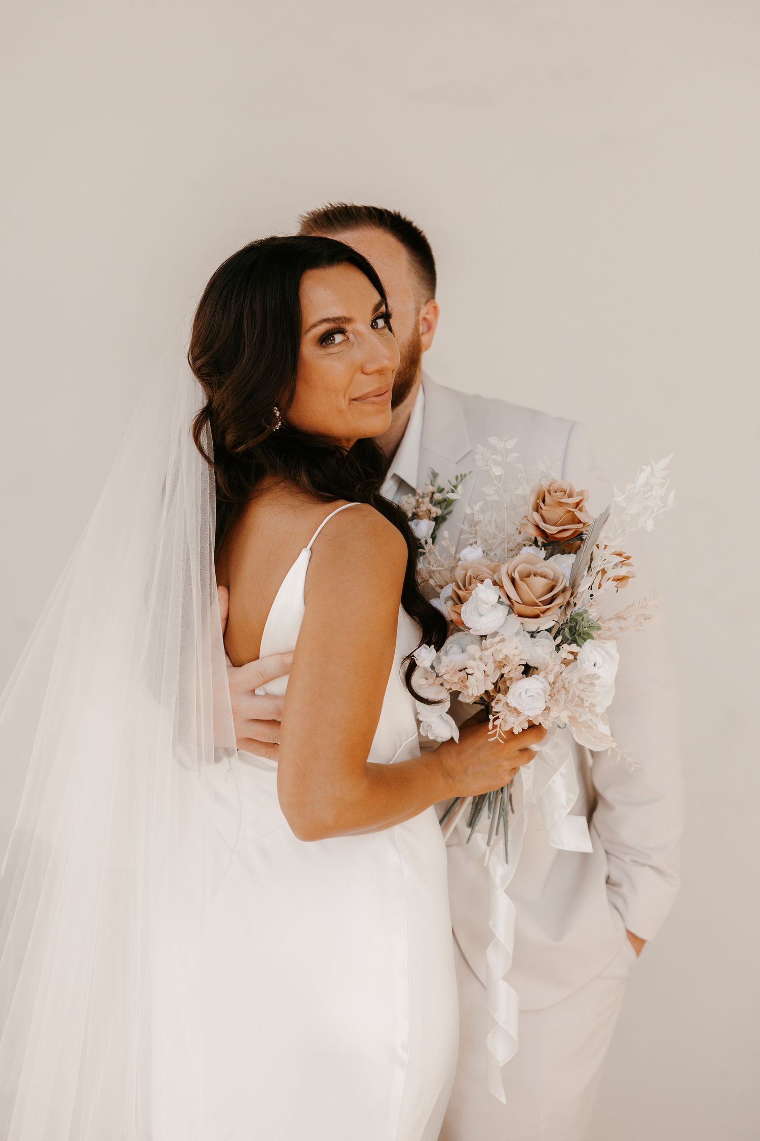 Bride holding boho bouquet while groom embraces her from behind against neutral white background.