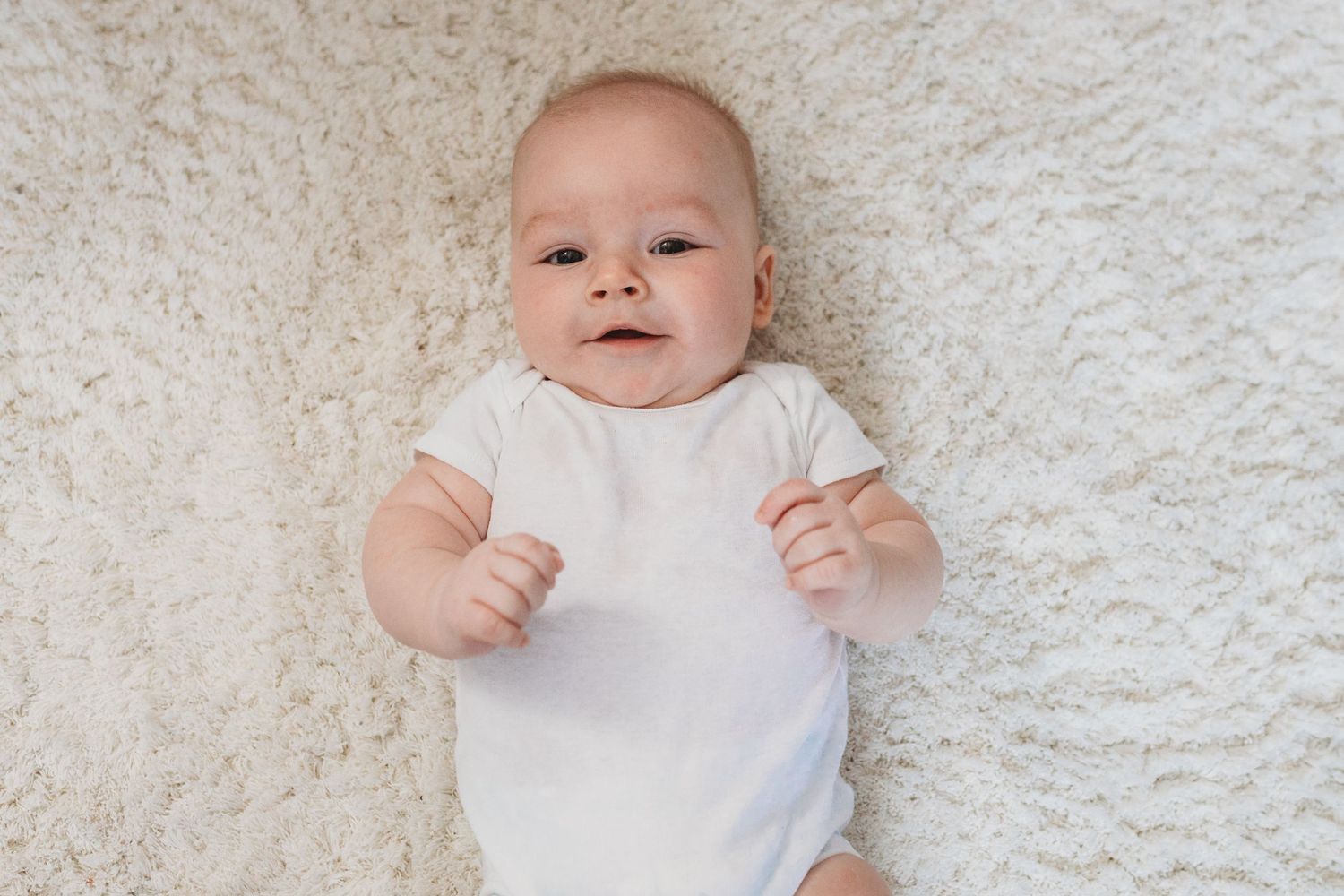 Baby wearing white onesie lies on textured white blanket during portrait session.