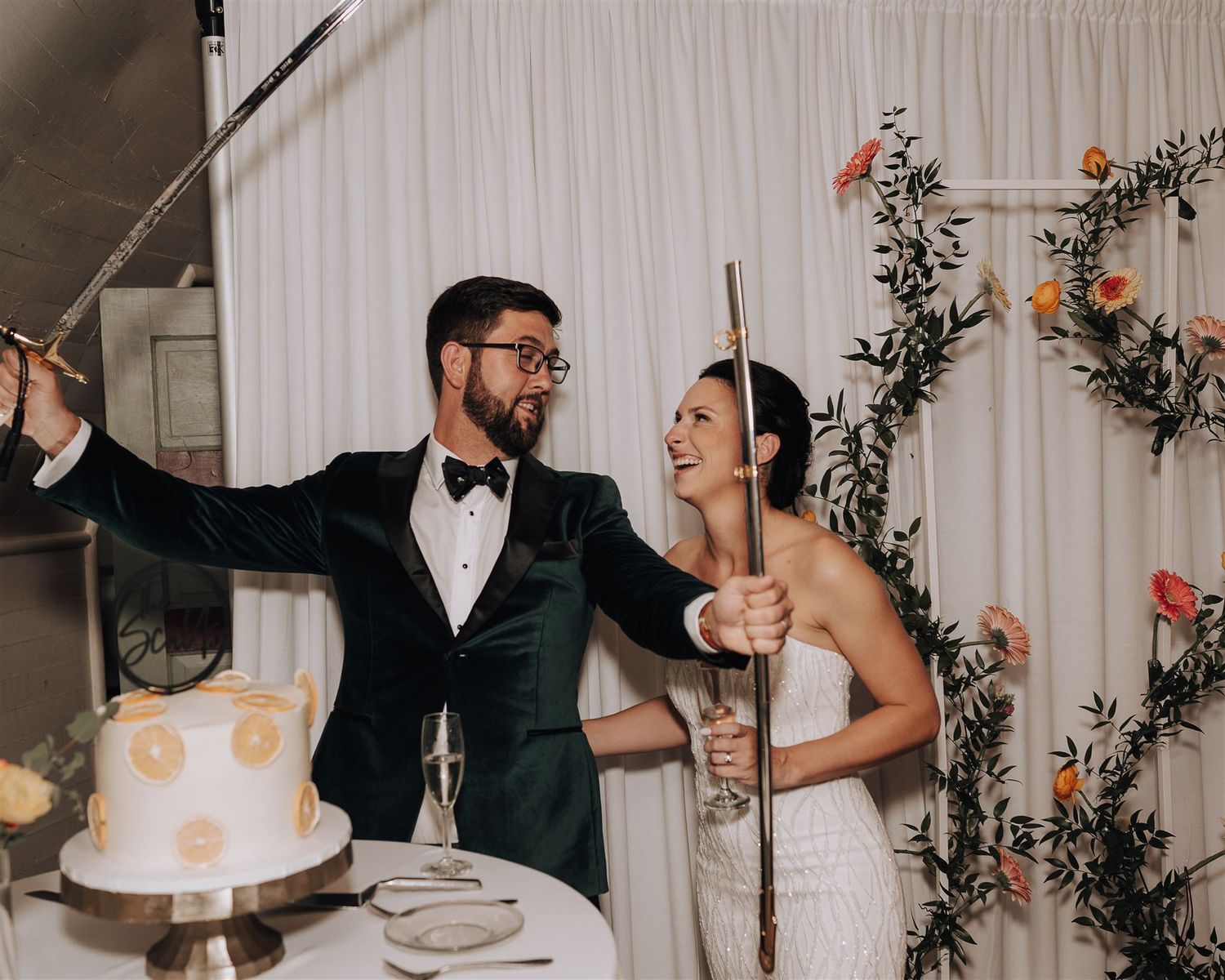 Couple celebrating joyfully next to their wedding cake with floral wall decoration.