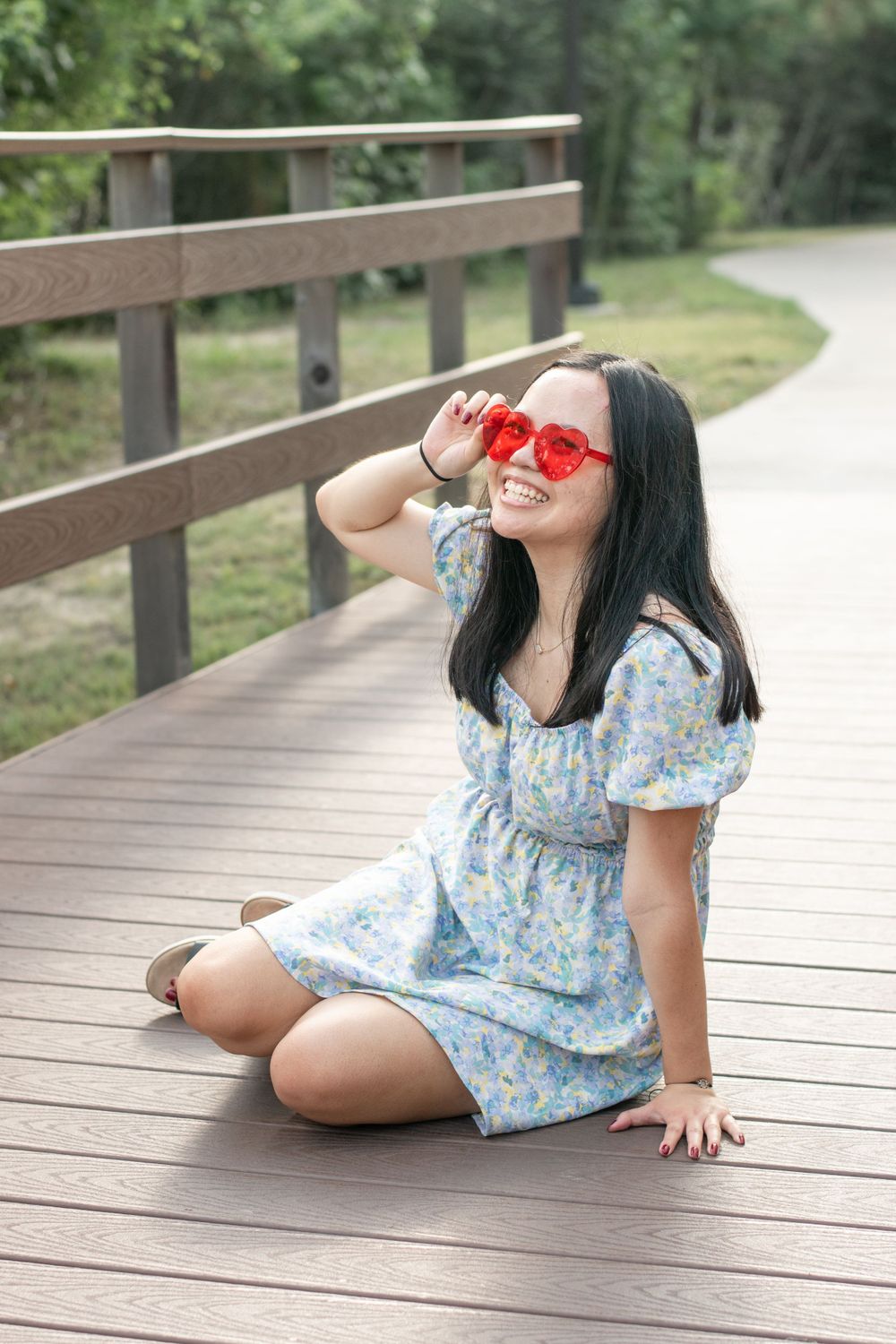 Person wearing heart-shaped red sunglasses sits playfully on wooden boardwalk.
