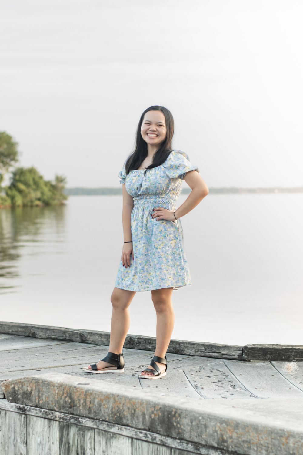 A person in a light blue floral dress poses on a concrete ledge by a waterfront.