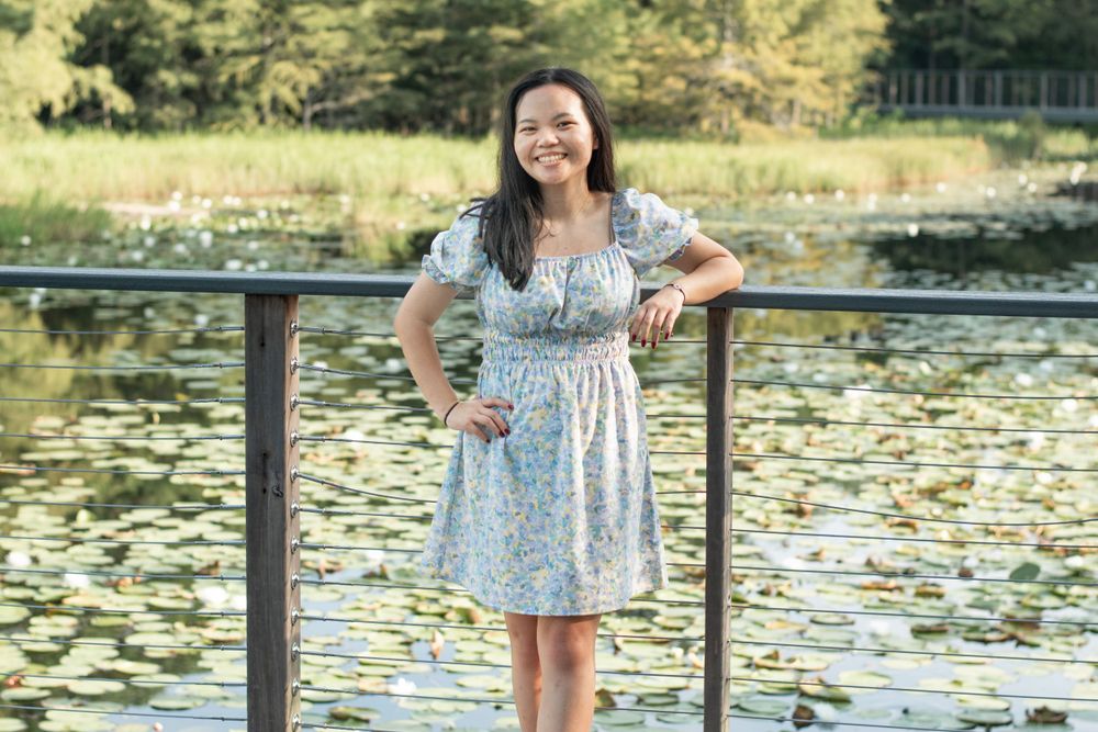 A person in a floral dress stands by a lake with lily pads visible in the water.
