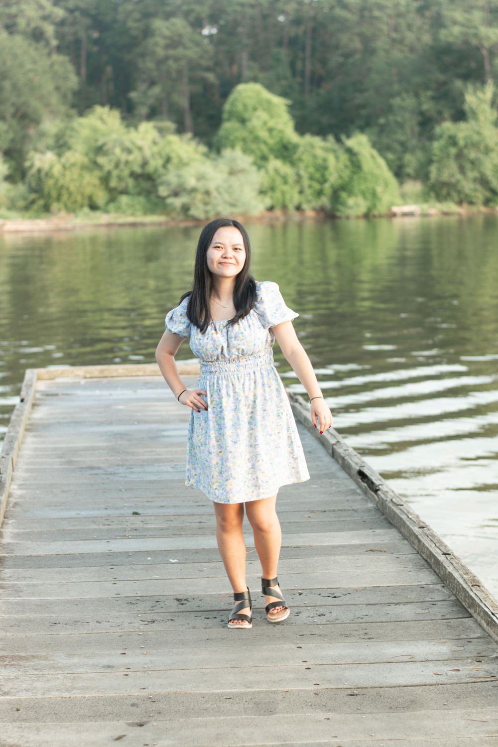 Person in a light summer dress poses on a wooden dock by a serene lake.