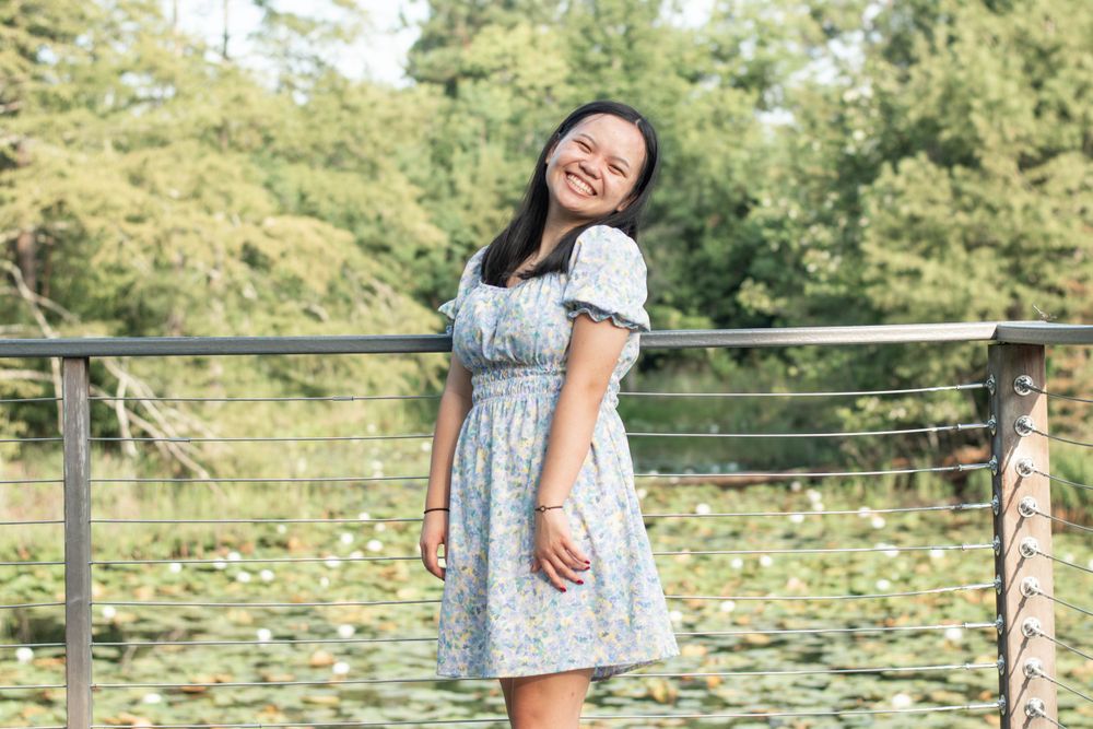 Person wearing a blue floral dress smiles while standing against a railing with greenery in background.