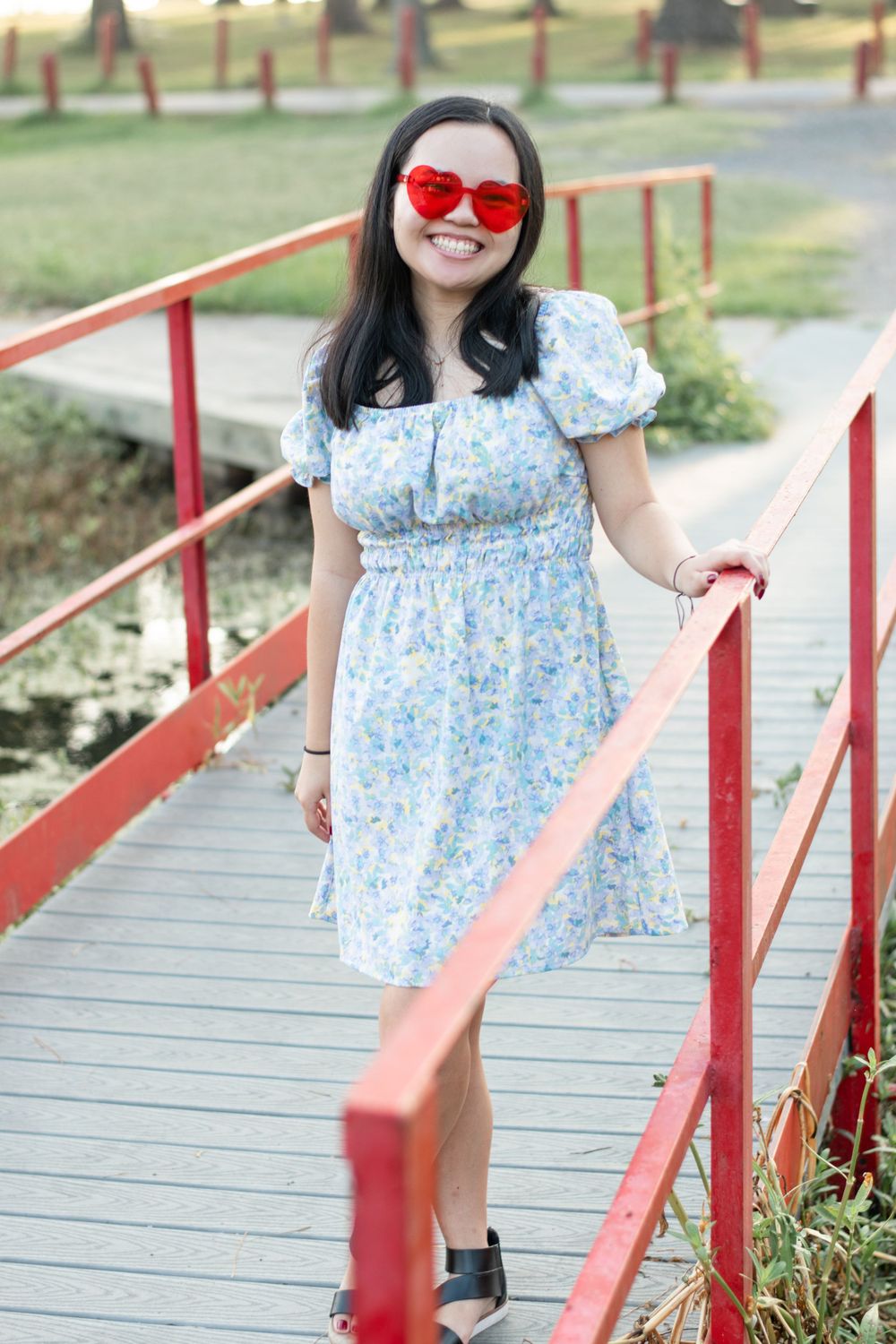 A person in a floral dress and red sunglasses stands on a red-railed bridge.