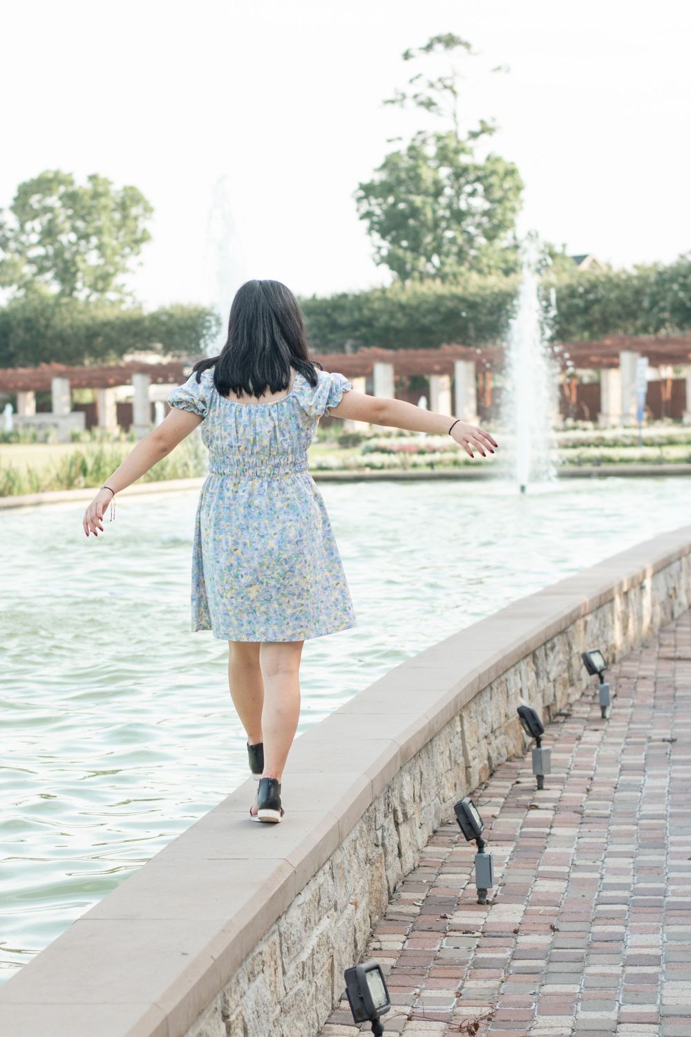 A person in a floral dress stands with arms outstretched near a fountain.