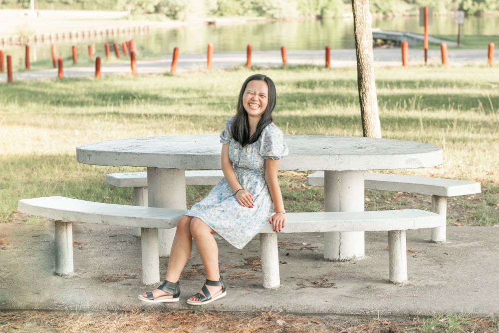 A person in a floral dress sits on a concrete bench in a park setting.