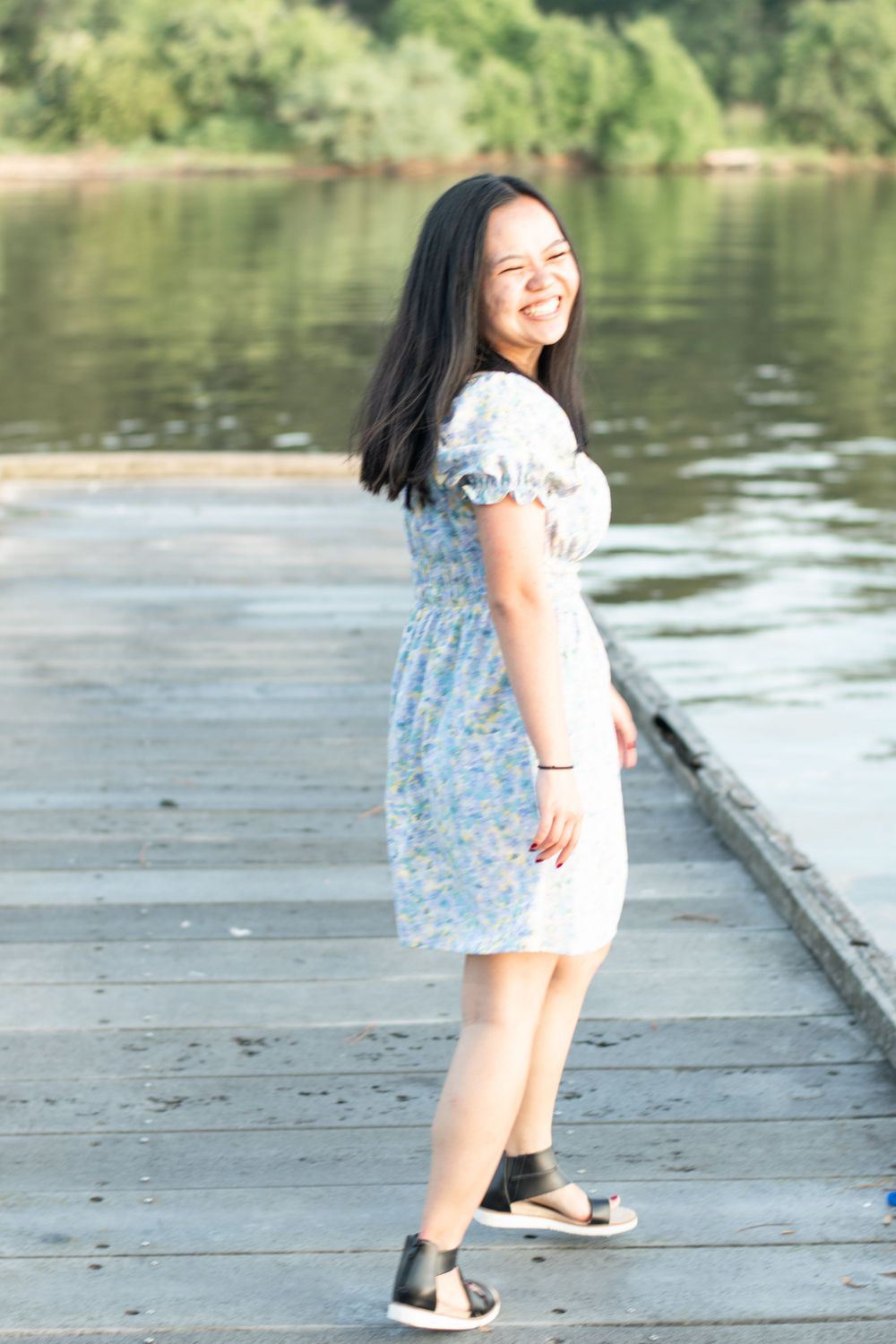 A person in a floral dress poses on a wooden dock with a lake and trees in the background.