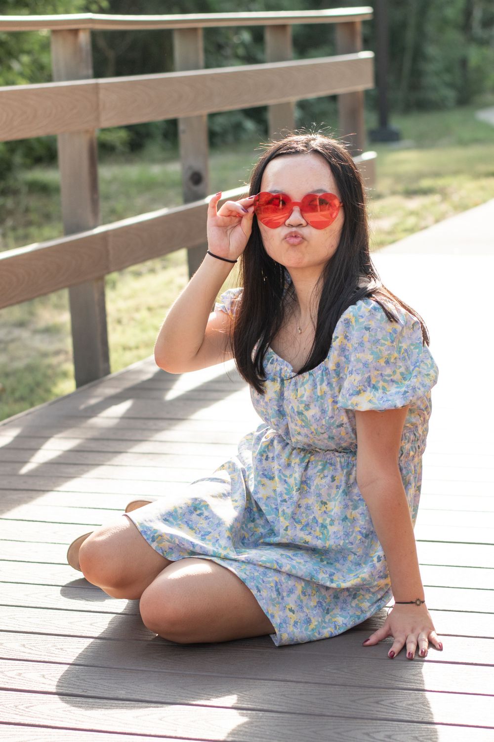 A person in a floral dress and red sunglasses makes a playful pose on a wooden deck.