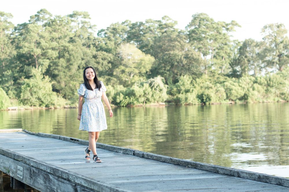 A person wearing a light blue dress walks along a wooden dock by a serene lake surrounded by trees.