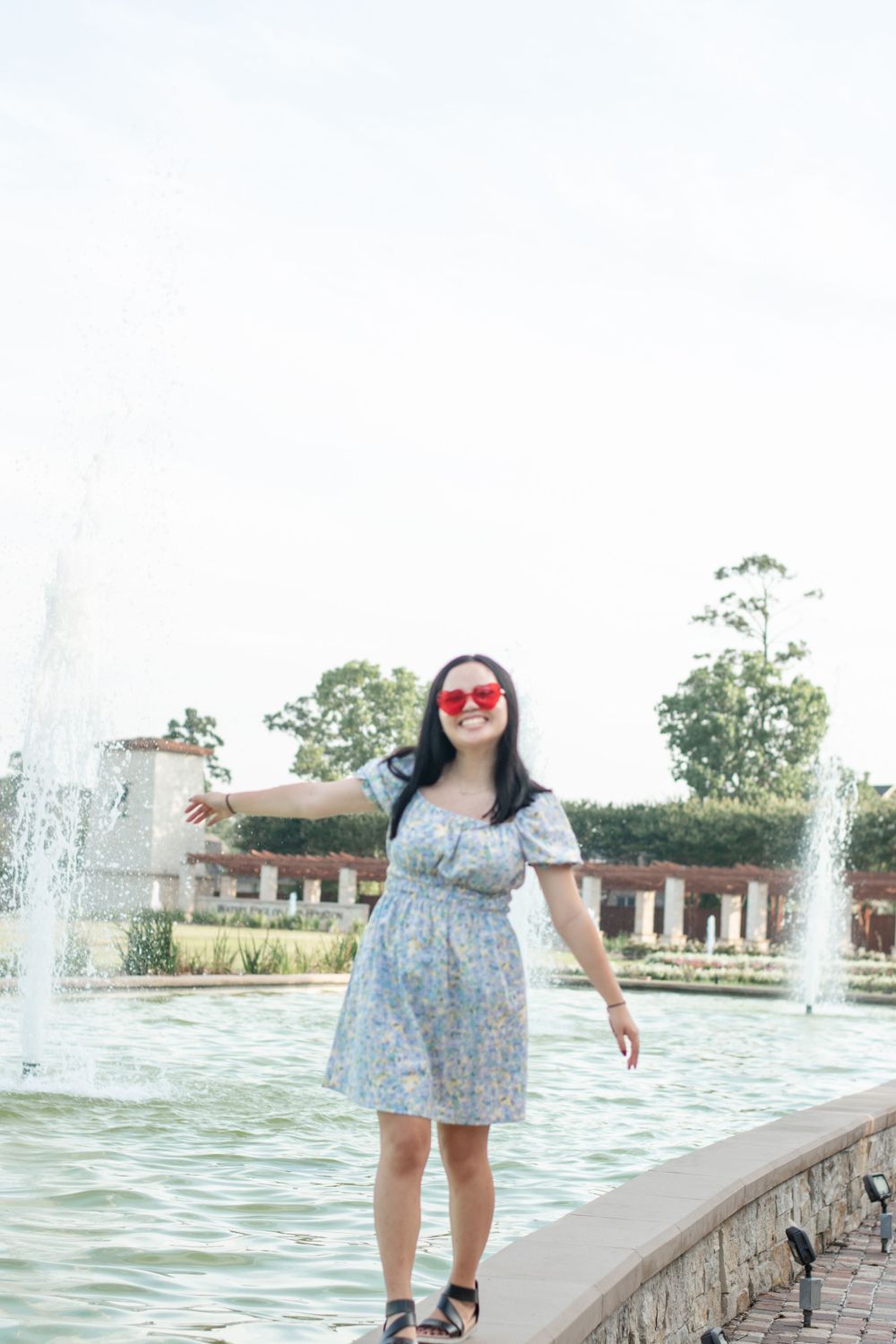 Person in a floral dress poses cheerfully by a fountain in a garden setting.